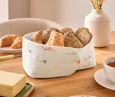 Na dřevěném stole stojí košík na chléb s různými druhy pečiva, včetně croissantů a housek, vedle šálku čaje, kostky másla a talíře s květinovým vzorem.