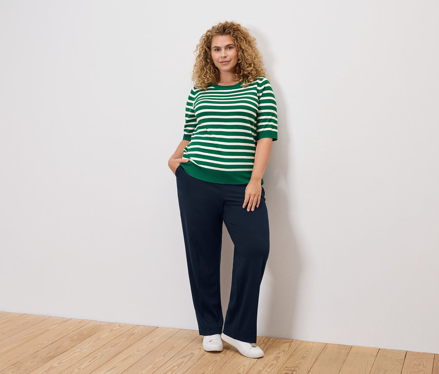 Žena s kudrnatými vlasy má na sobě zeleno-bílý pruhovaný top, tmavě modré kalhoty a bílé boty.