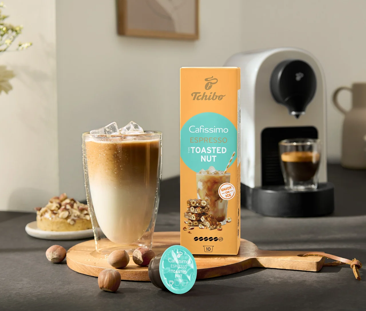 Sklenice ledové kávy stojí vedle balení Cafissimo Flavoured Espresso – Toasted Nut a kávovaru Cafissimo pure+, white.