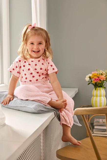 Usmívající se dívka sedí v 2 Kinder-T-Shirts a Kinder-Musselin-Hose na polštáři. Váza stojí na židli.