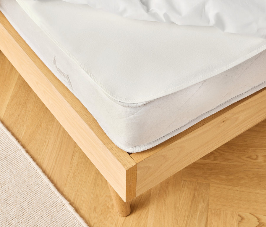 Detailní pohled na postel s bílou matrací a ložním prádlem na světlé dřevěné podlaze, vedle béžový koberec.