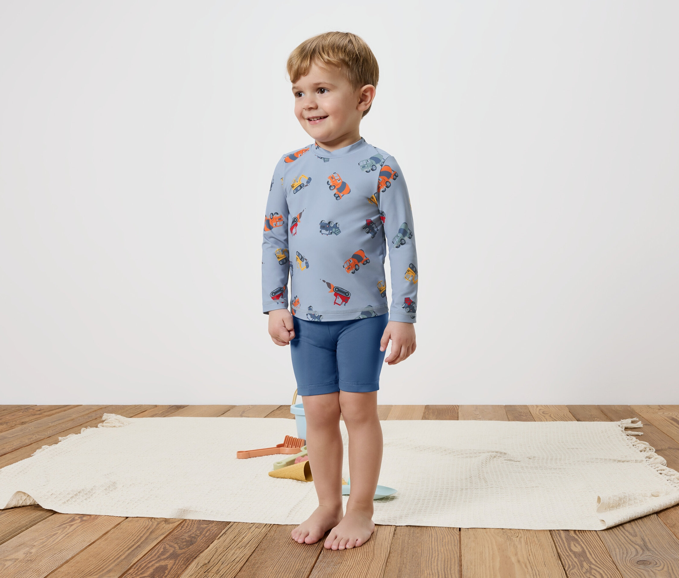 Malý chlapec stojí bosý na koberci a má na sobě modrý top s dlouhým rukávem s barevnými vozidly a modré šortky.