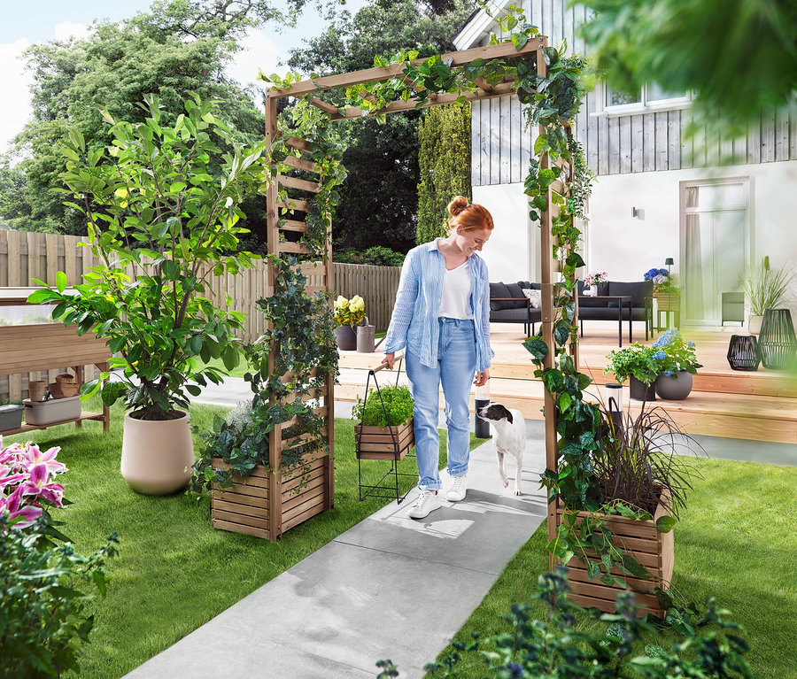 Žena prochází se psem zahradním obloukem porostlým rostlinami. Nese květináč.