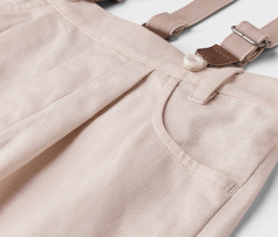 Detailní záběr světle béžových kalhot s šlemi, knoflíkem a kapsou.