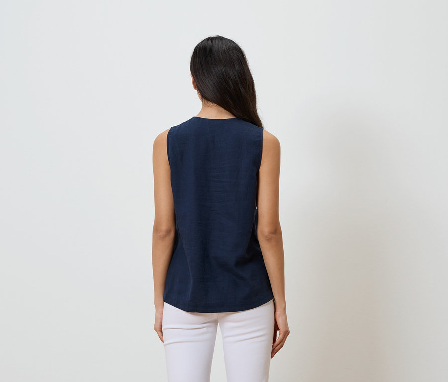 Žena s tmavými vlasy, viděná zezadu, má na sobě tmavě modrý top bez rukávů a bílé kalhoty na bílém pozadí.