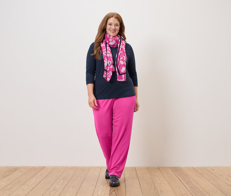 Žena s dlouhými rusými vlasy stojí před bílou zdí a má na sobě tmavě modrou halenku, růžové kalhoty a růžový šátek s květinami.