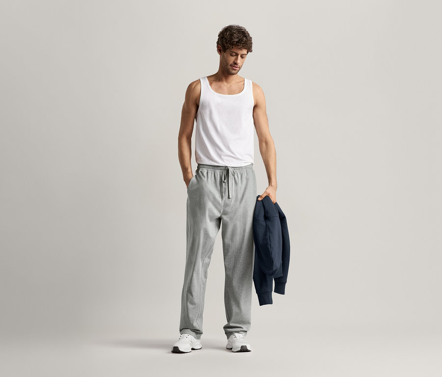 Muž v bílém tílku a šedých pohodlných kalhotách, s jednou rukou v kapse a tmavě modrou bundou přehozenou přes rameno.