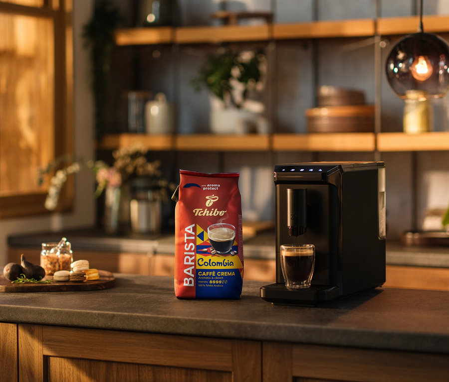 Na kuchyňské lince stojí kávovar, šálek kávy a balení BARISTA Origins Colombia − 1 kg zrnkové kávy.