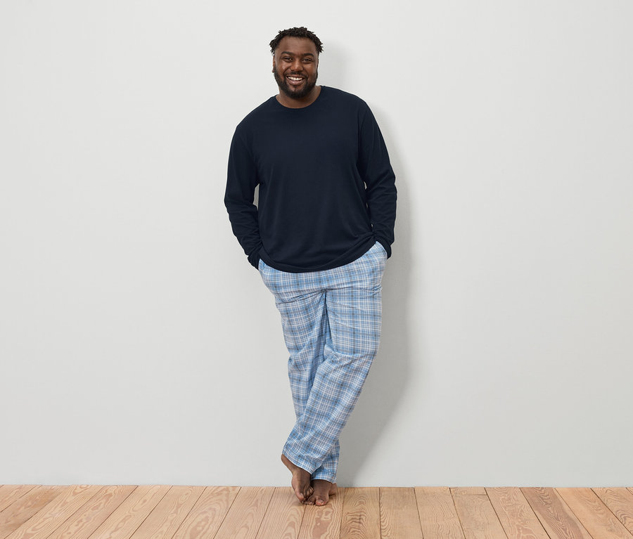 Muž tmavé pleti s vousy v tmavě modré košili s dlouhým rukávem a modrobílých kostkovaných pyžamových kalhotách stojí bosý na dřevěné podlaze před bílou stěnou.