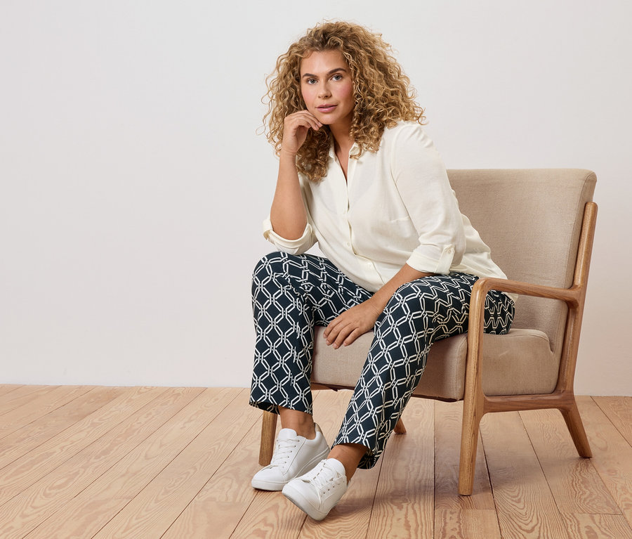 Žena s kudrnatými vlasy sedí v křesle ve vzorovaných kalhotách a bílých teniskách.