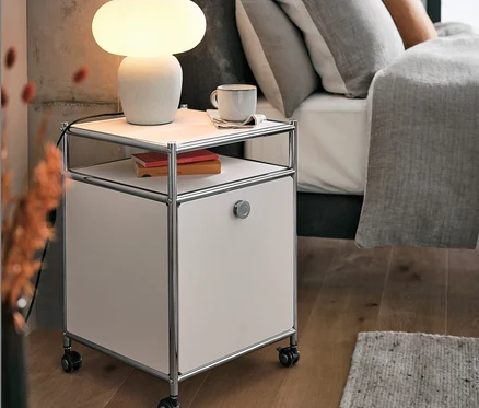 Stolní lampa se skleněným stínítkem stojí na kovovém kontejneru na kolečkách CN3 s výklopnými dvířky, bílý, vedle postele.