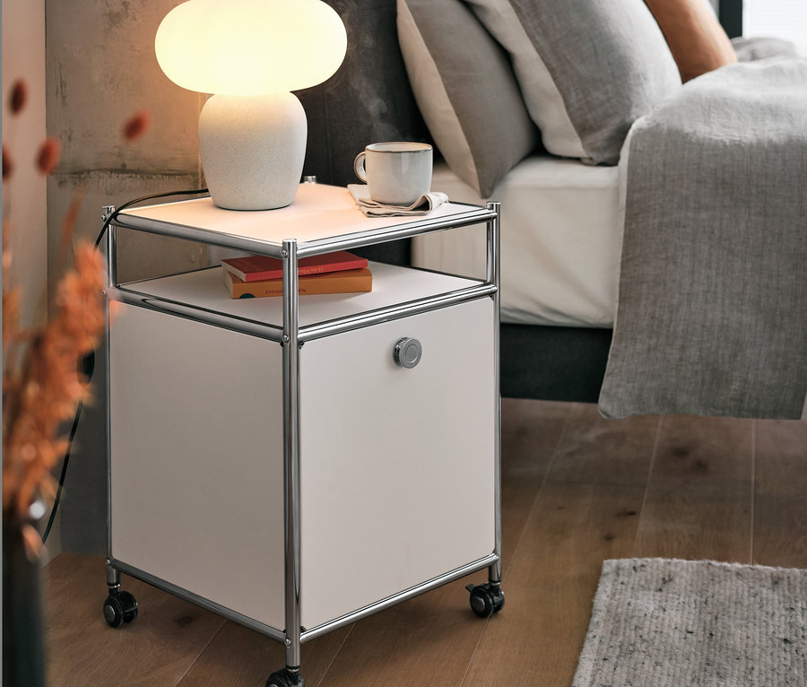 Stolní lampa se skleněným stínítkem stojí na kovovém kontejneru na kolečkách CN3 s výklopnými dvířky, bílý, vedle postele.