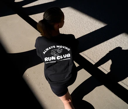 Žena zezadu v černé mikině s nápisem „Always Moving Run Club“ a krátkých antracitových sportovních legínách »ActivePower« na cestě se stíny.