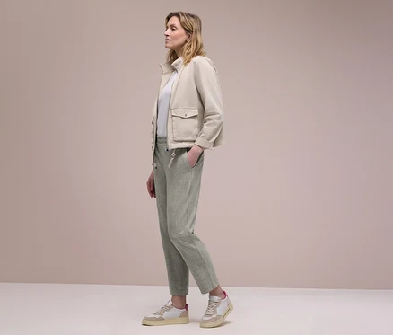 Žena z profilu v béžové bundě, bílém topu a světle zelených kalhotách joggpant Street One, s jednou rukou v kapse. Má na sobě béžovo-bílé tenisky.