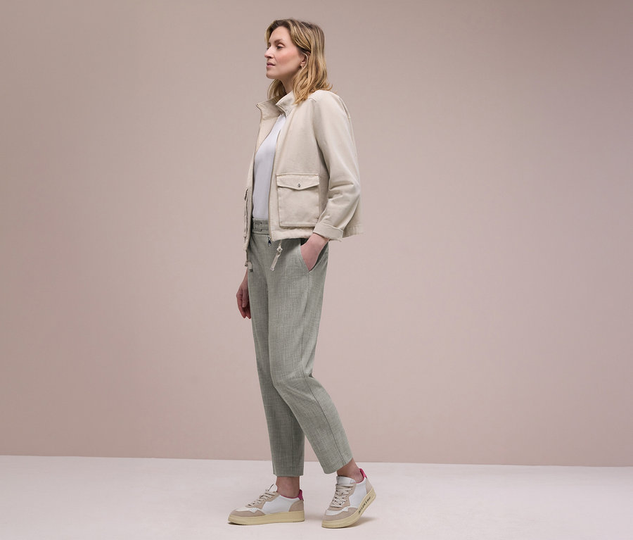 Žena z profilu v béžové bundě, bílém topu a světle zelených kalhotách joggpant Street One, s jednou rukou v kapse. Má na sobě béžovo-bílé tenisky.