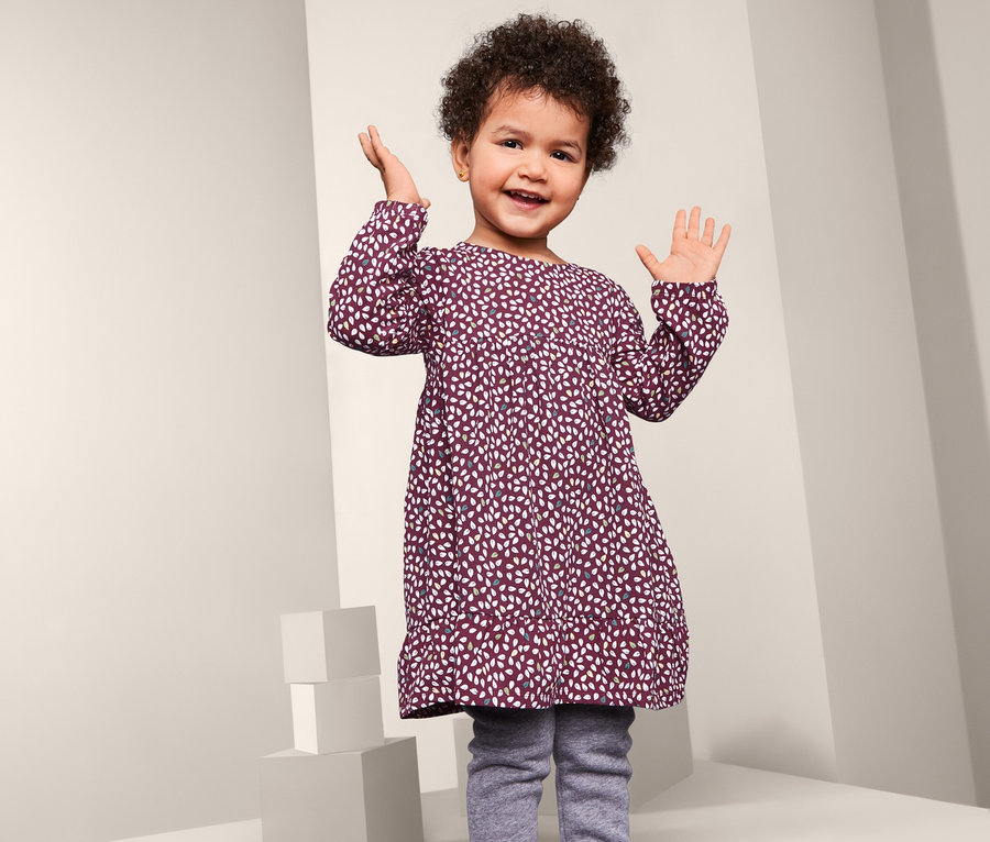 Usmívající se holčička pózuje v džerzejových šatech s dlouhým rukávem a vzorem.