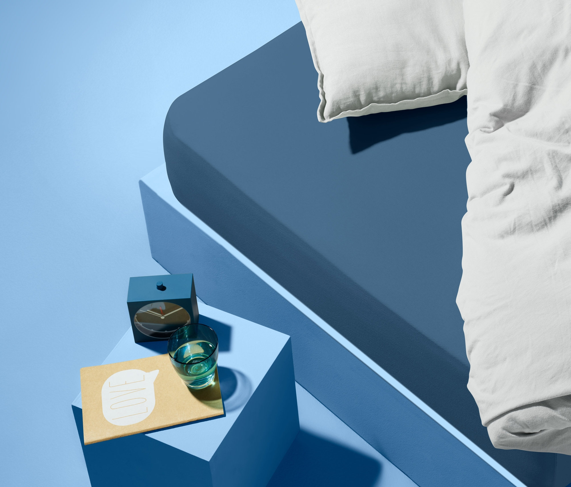 Modrá postel s bílým polštářem a peřinou. Vedle postele je modrý noční stolek s budíkem, sklenicí vody a poznámkovým blokem.