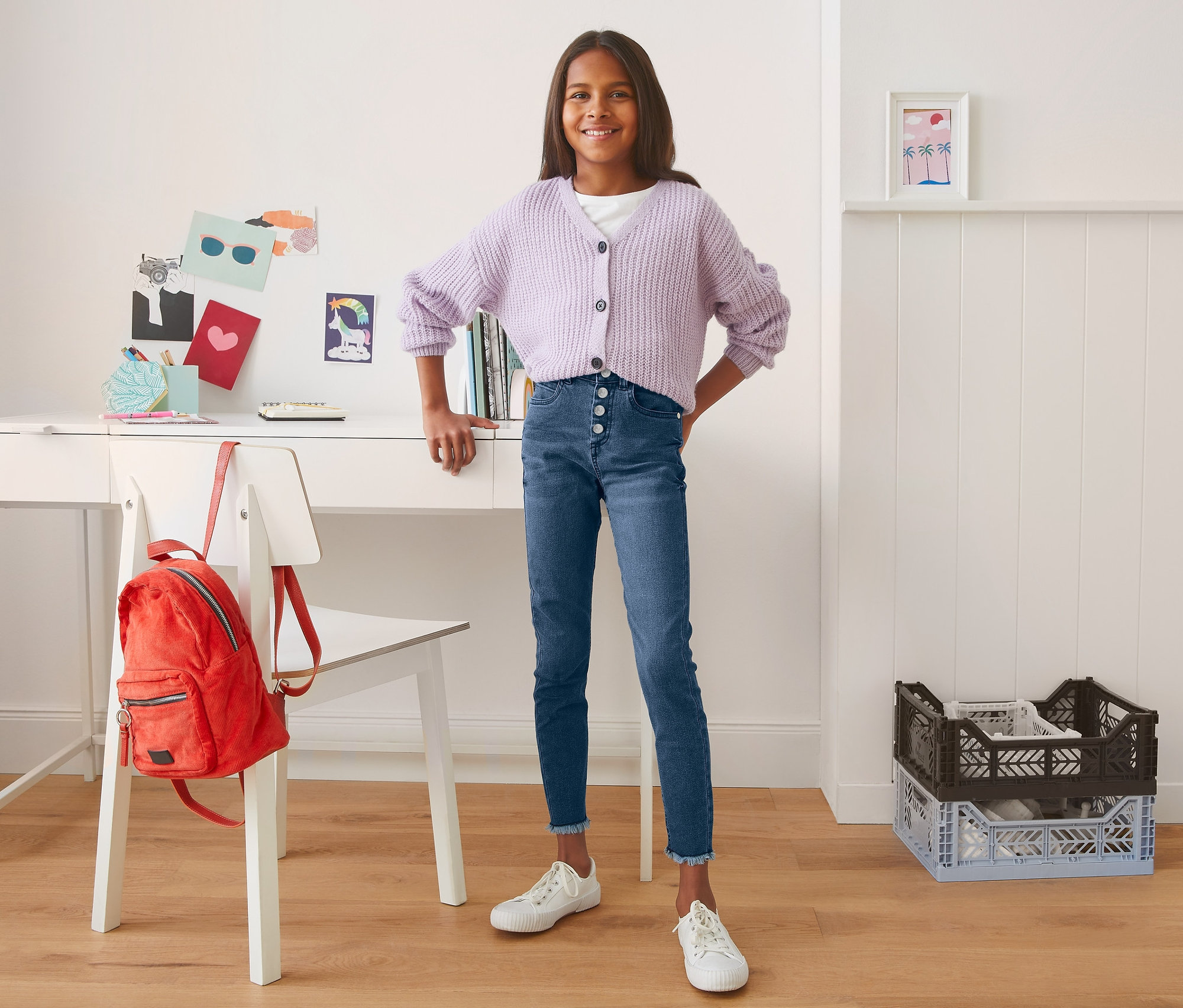 Usmívající se dospívající dívka pózuje v modrých džínách s vysokým pasem a fialovém svetru před stolem.