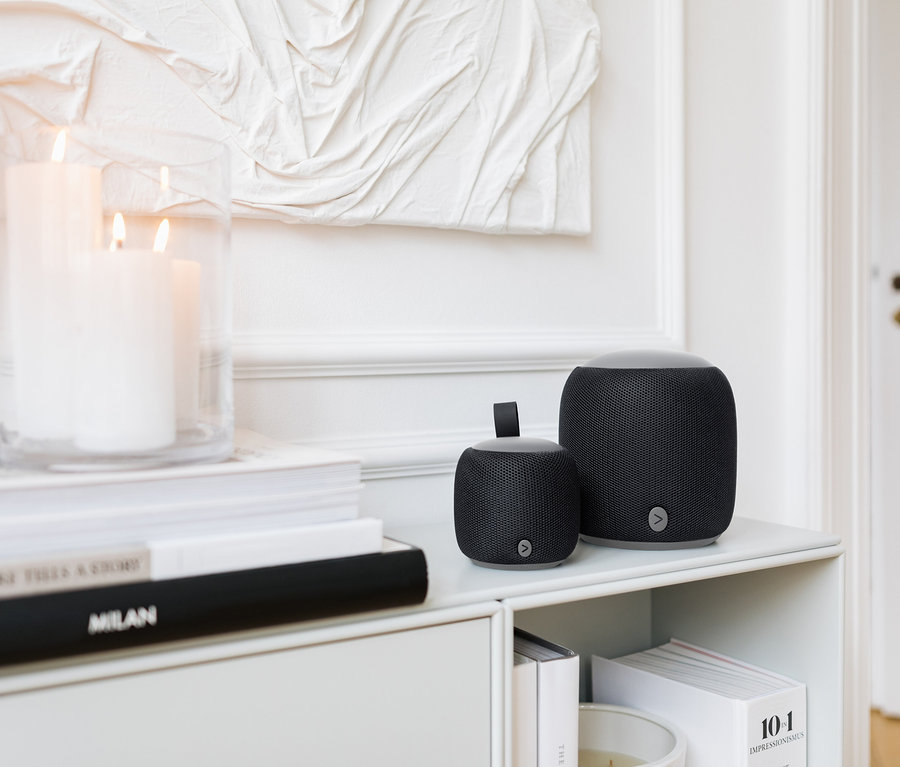 Dva černé reproduktory Bluetooth Fabric různých velikostí stojí na bílé polici vedle svíček a knih.