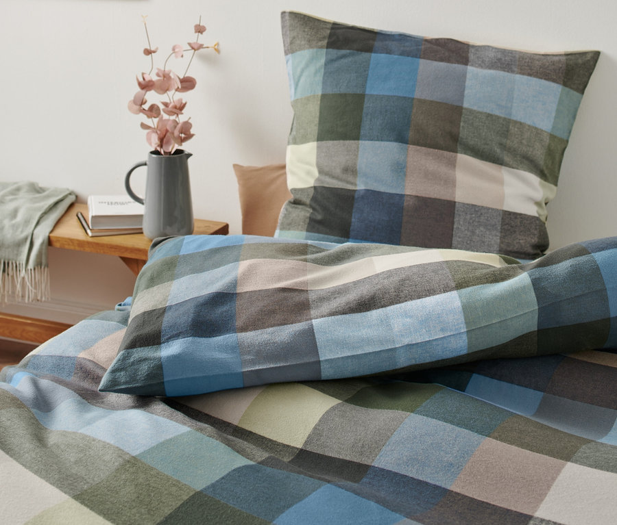 Kostkované flanelové povlečení, standardní velikosti, leží na posteli vedle vázy s květinami.
