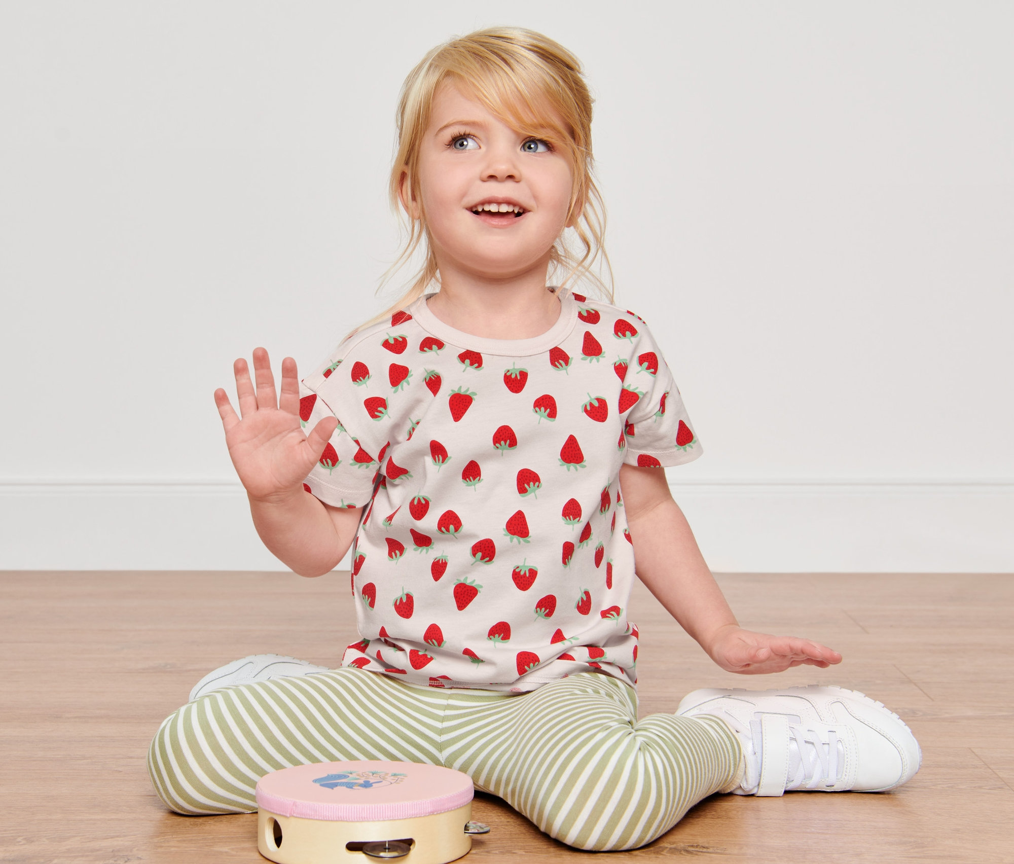 Dívka sedí na podlaze a má na sobě růžové tričko s jahodami a zelenobílé pruhované kalhoty. V popředí leží dětská trička, 3 ks, růžová, zelená/bílá a světle modrá.