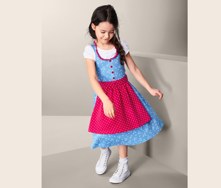 Dívka v modrých žerzejových dirndl šatech s červenou zástěrou s bílými puntíky a bílých teniskách.