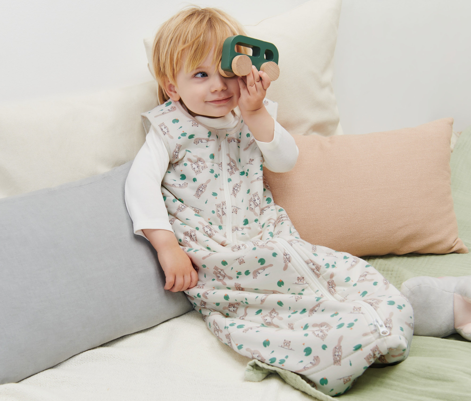 Dítě leží v spacím pytli s vydrami a hraje si s dřevěným autíčkem.