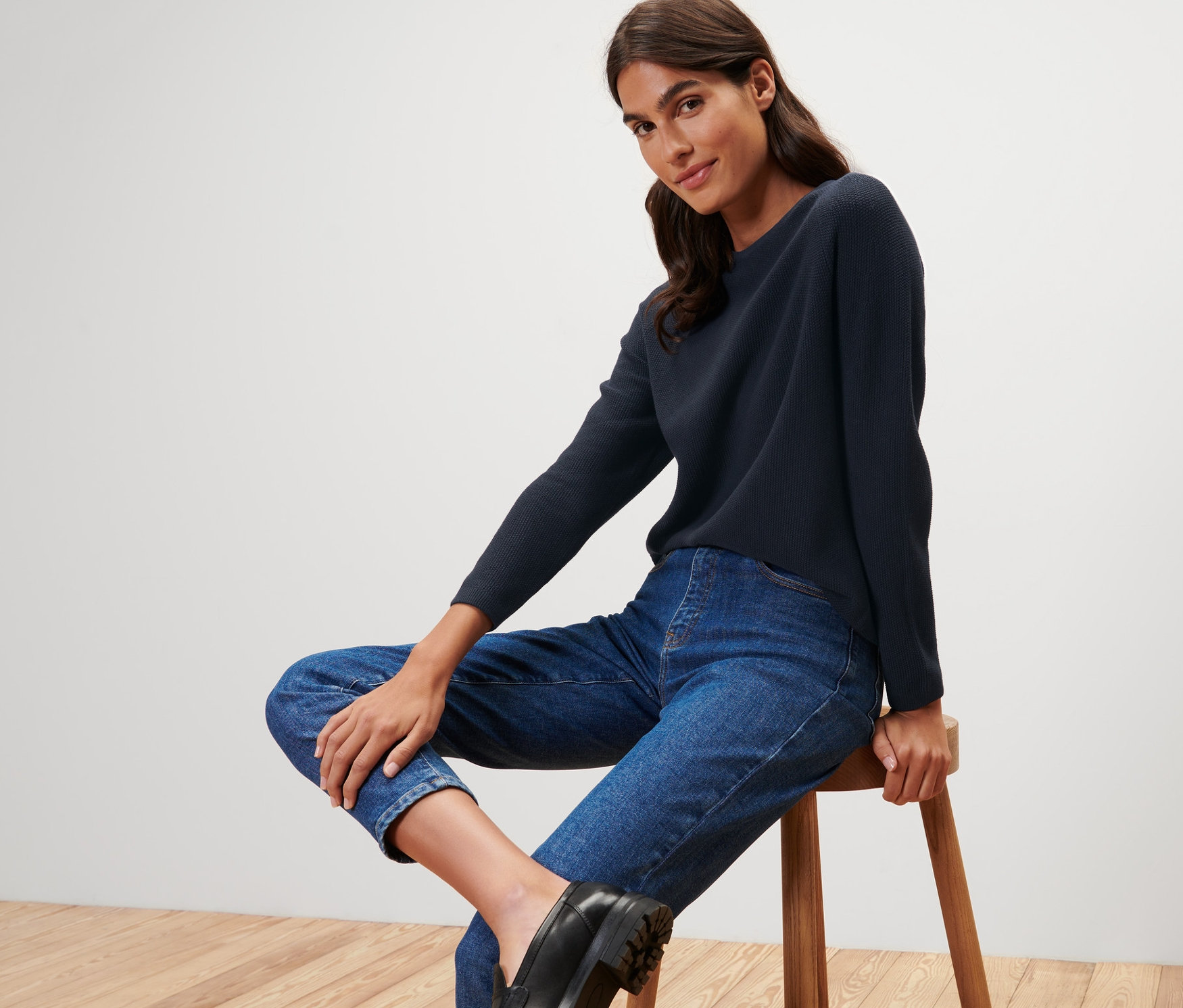 Žena sedí na stoličce a má na sobě tmavě modrý pletený svetr.