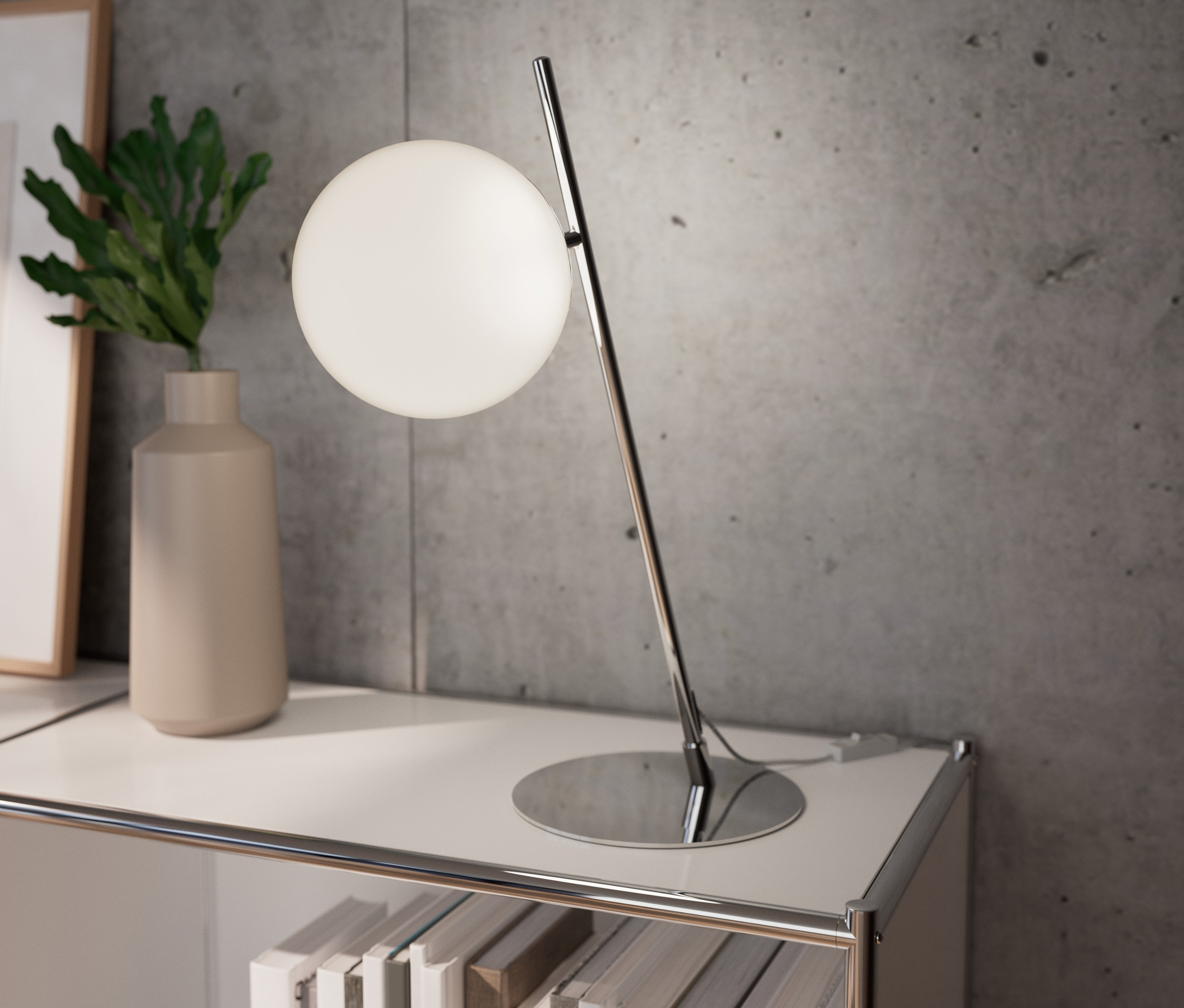 Chromovaná kovová stolní lampa stojí na bílé polici vedle vázy s rostlinou.
