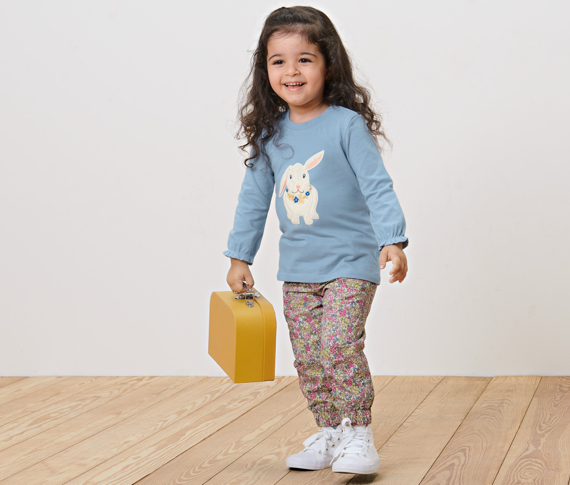 Usmívající se holčička drží žlutý kufřík, má na sobě modré tričko s dlouhým rukávem s králíčkem a květinové kalhoty.