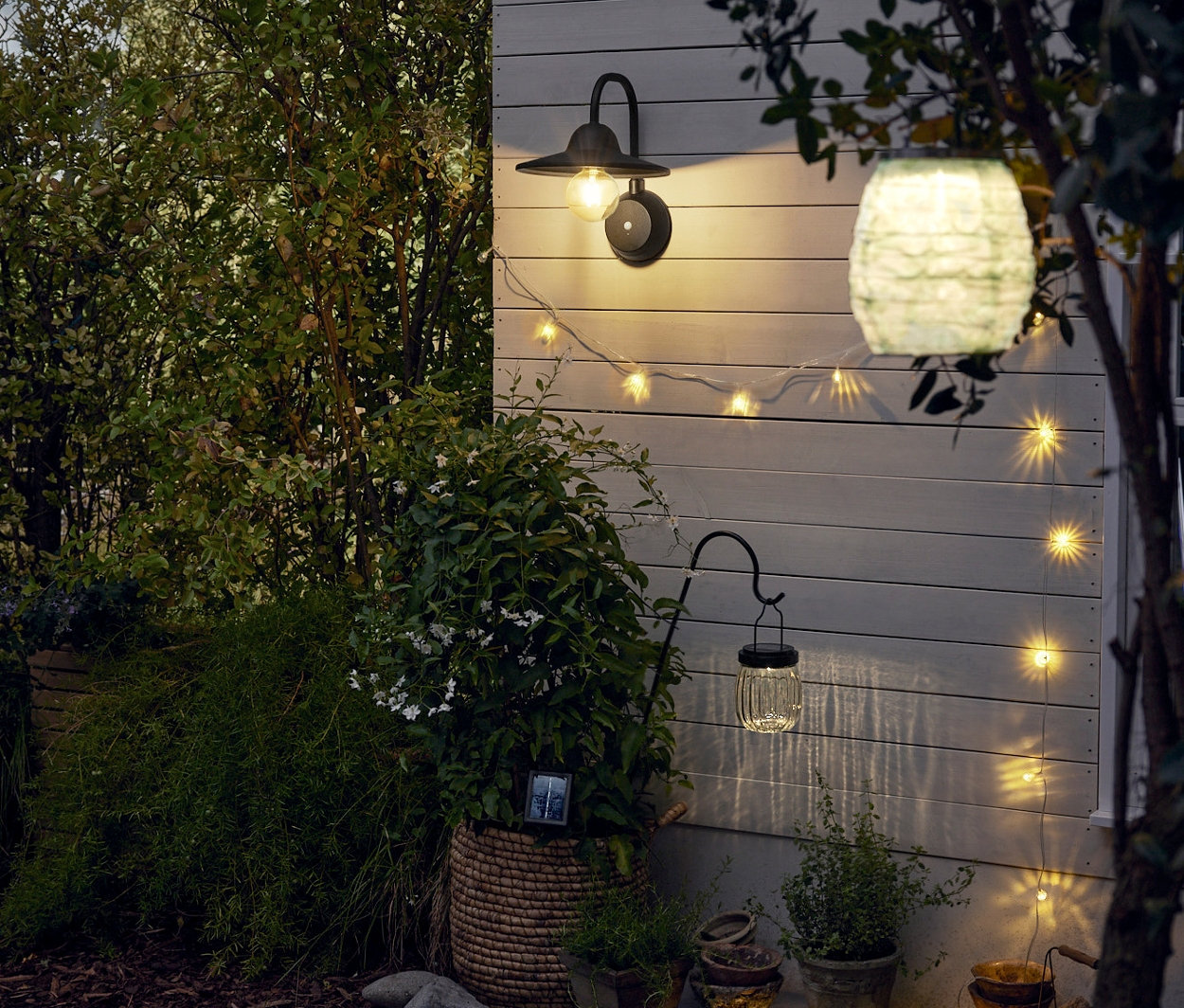 Na zdi visí LED nástěnné svítidlo s přídavnou výměnnou baterií. Vedle něj je solární světelný řetěz. V popředí je solární lampión a solární svítidlo s bodcem.