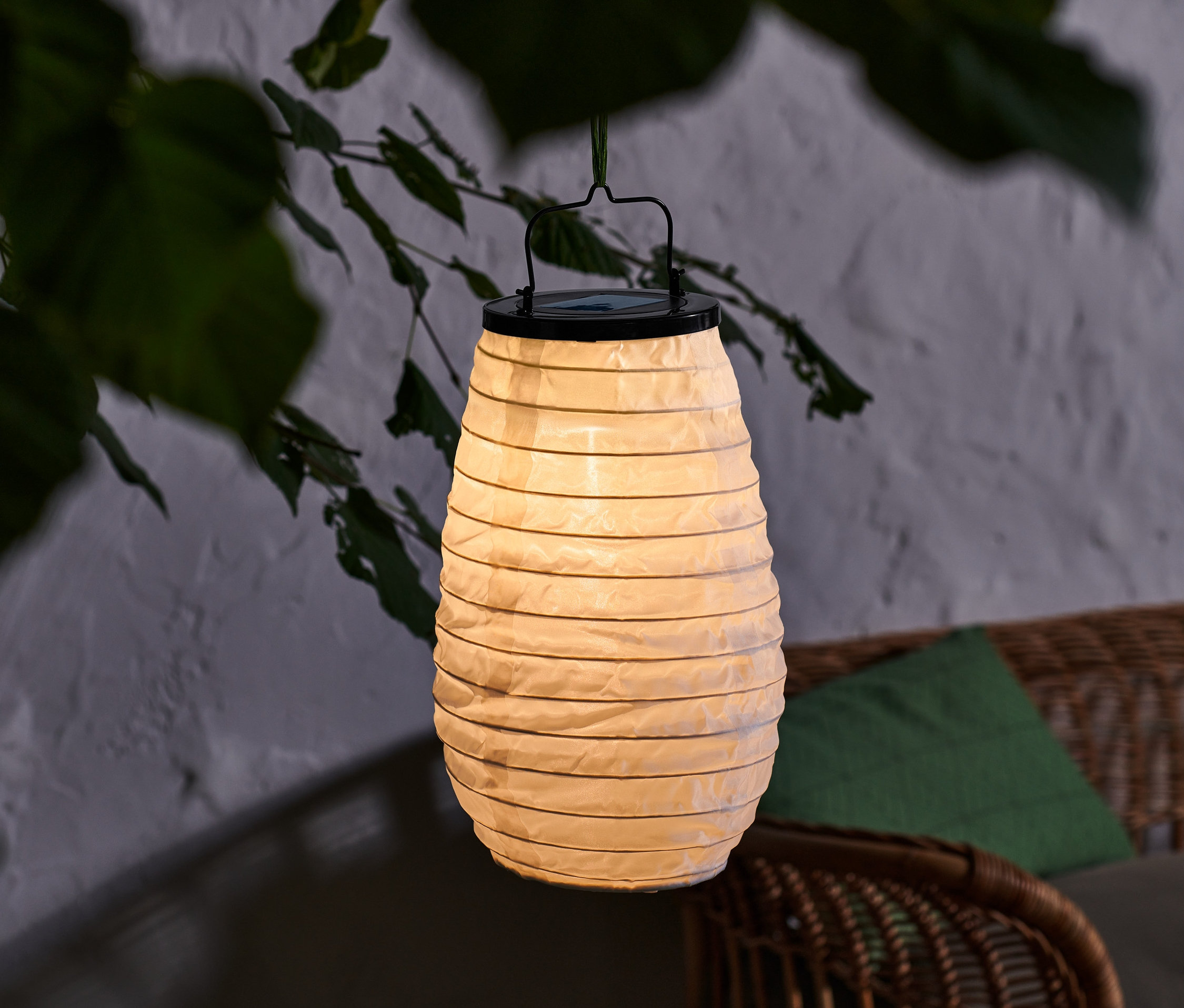 Zavěšený solární lampión s černým držadlem, umístěný venku mezi větvemi stromu.
