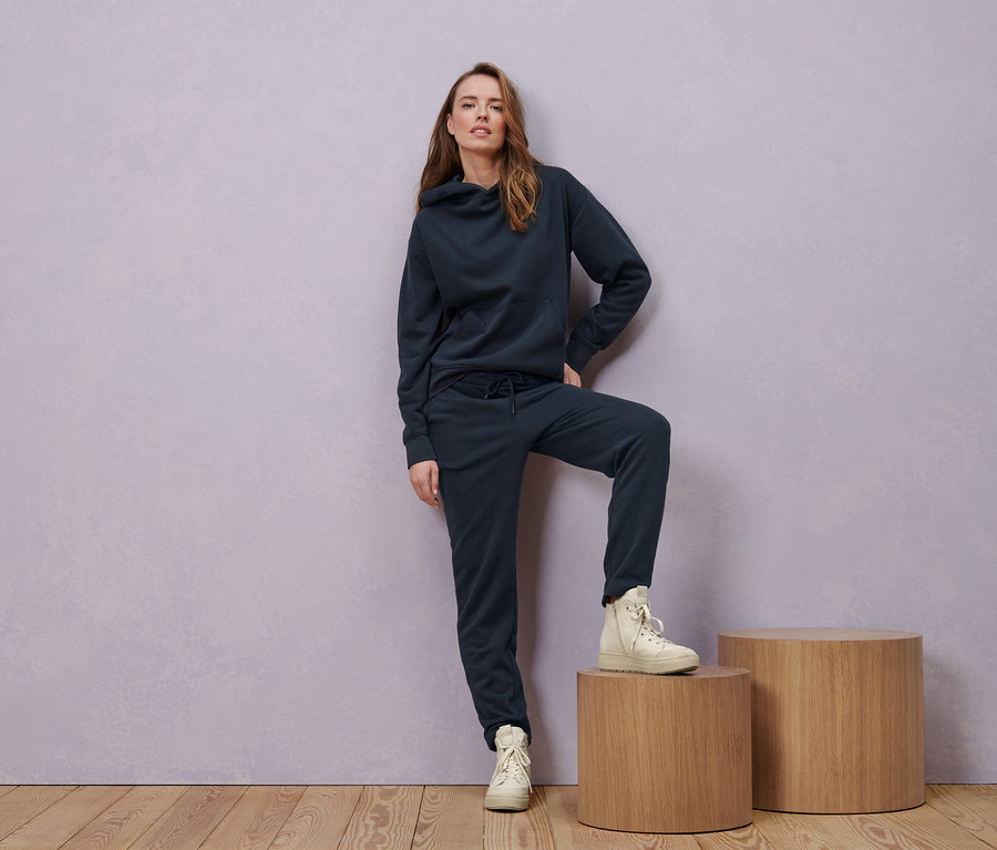 Žena pózuje v námořnicky modré mikině s kapucí, teplákových kalhotách a kožených botách sneaker.