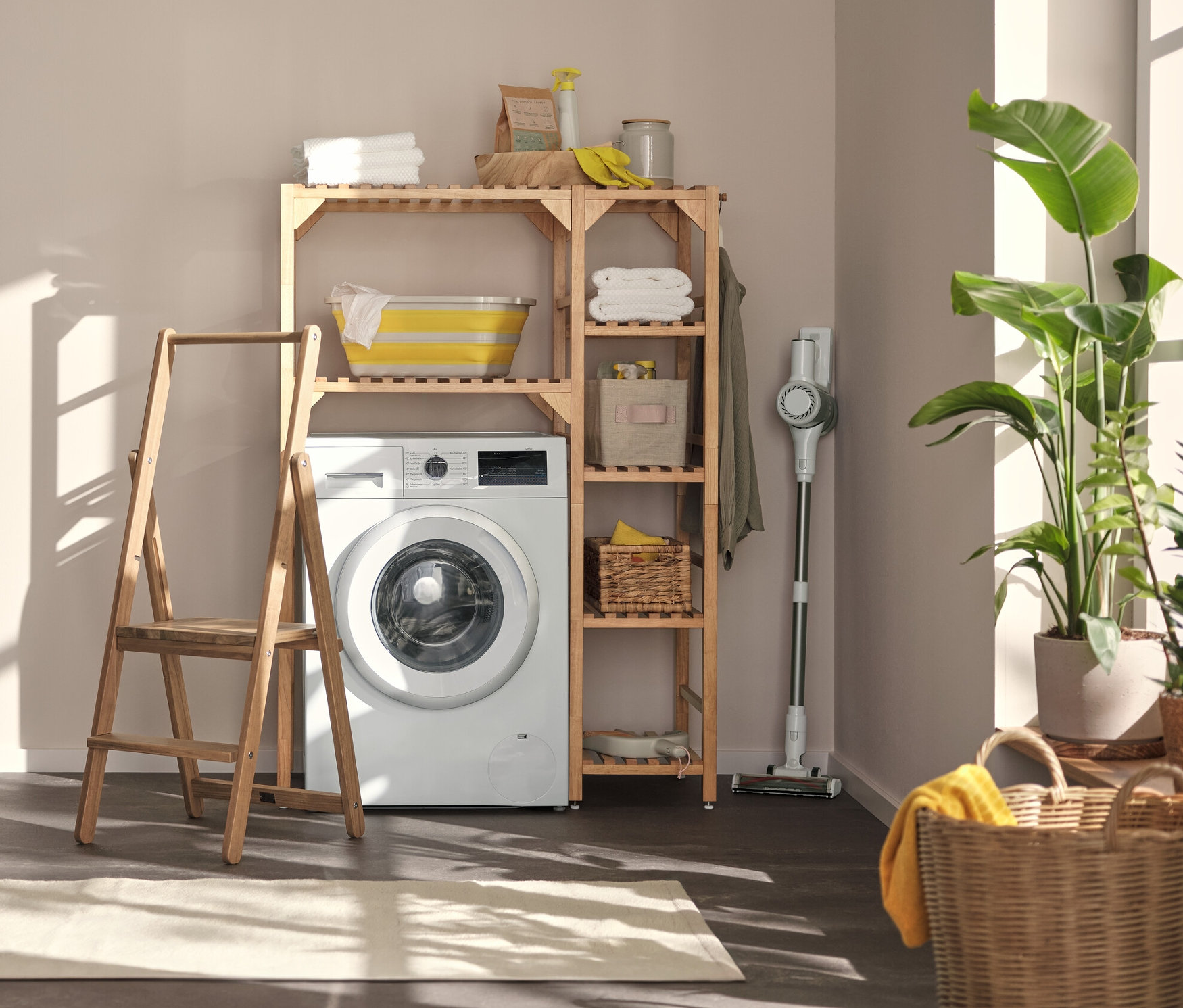 V prádelně stojí pračka, nad ní regál na pračku s bočním prvkem. Vlevo je nástěnný regál s flexibilními háčky a policí a schůdky z masivního dřeva.