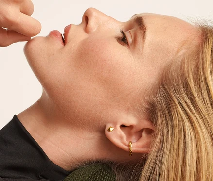 Žena leží na zeleném křesle a dotýká se rtu. Má stříbrné náušnice 925 pyramida a stříbrné náušnice 925 ear cuff pyramida.