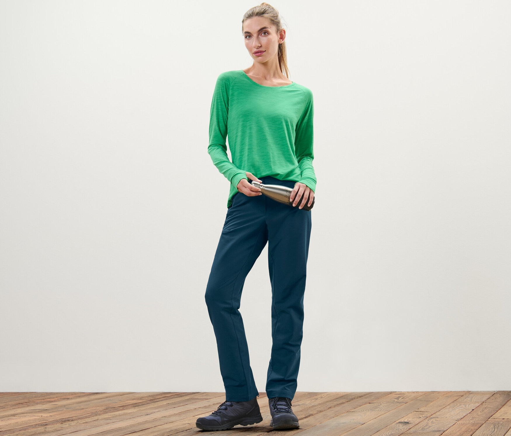 Žena v zeleném tričku drží stříbrnou láhev a má na sobě softshellové kalhoty.