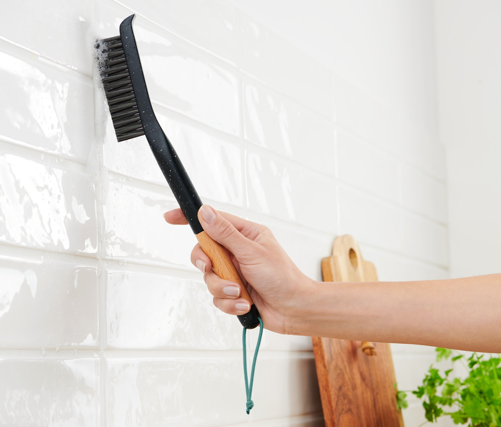 Žena čistí bílé dlaždice dlouhým kartáčem na spáry.