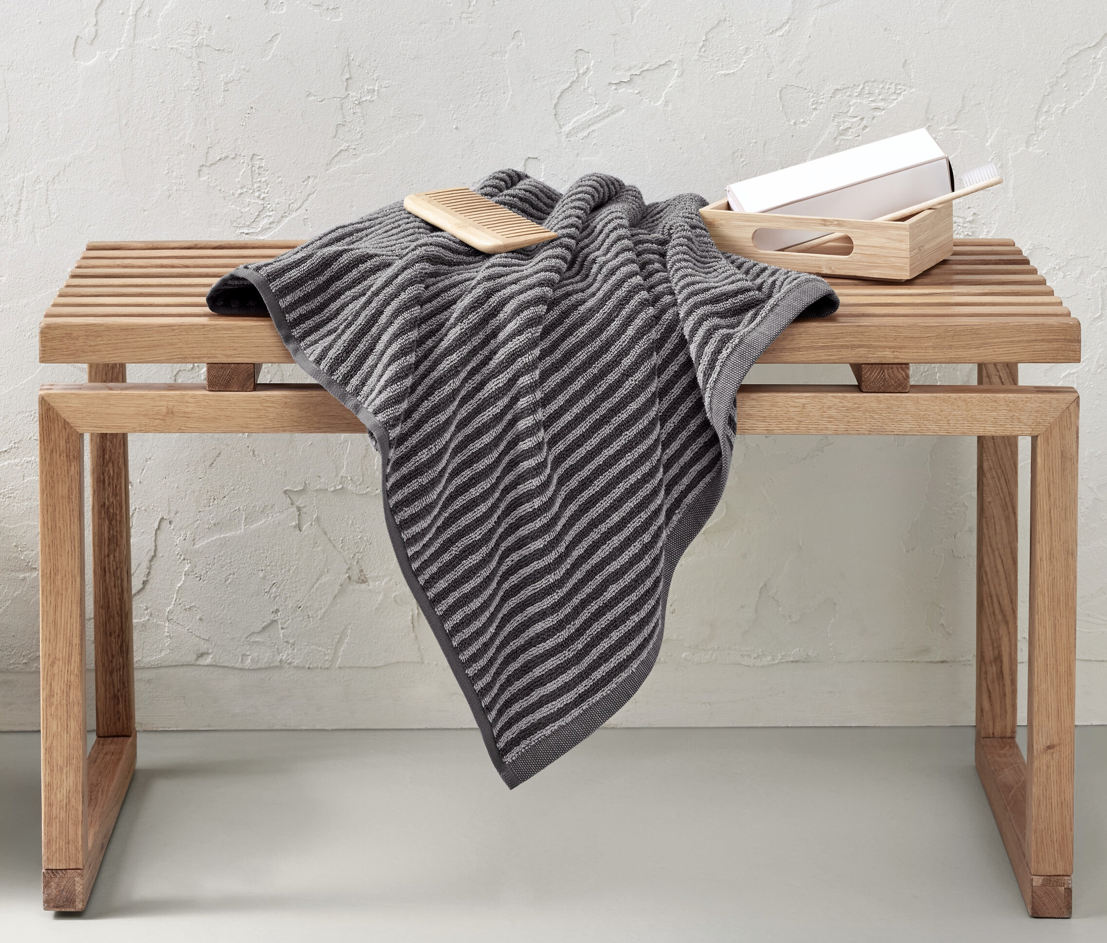 Na dřevěné lavici leží kvalitní šedá osuška. Vedle lavice stojí proutěný koš.