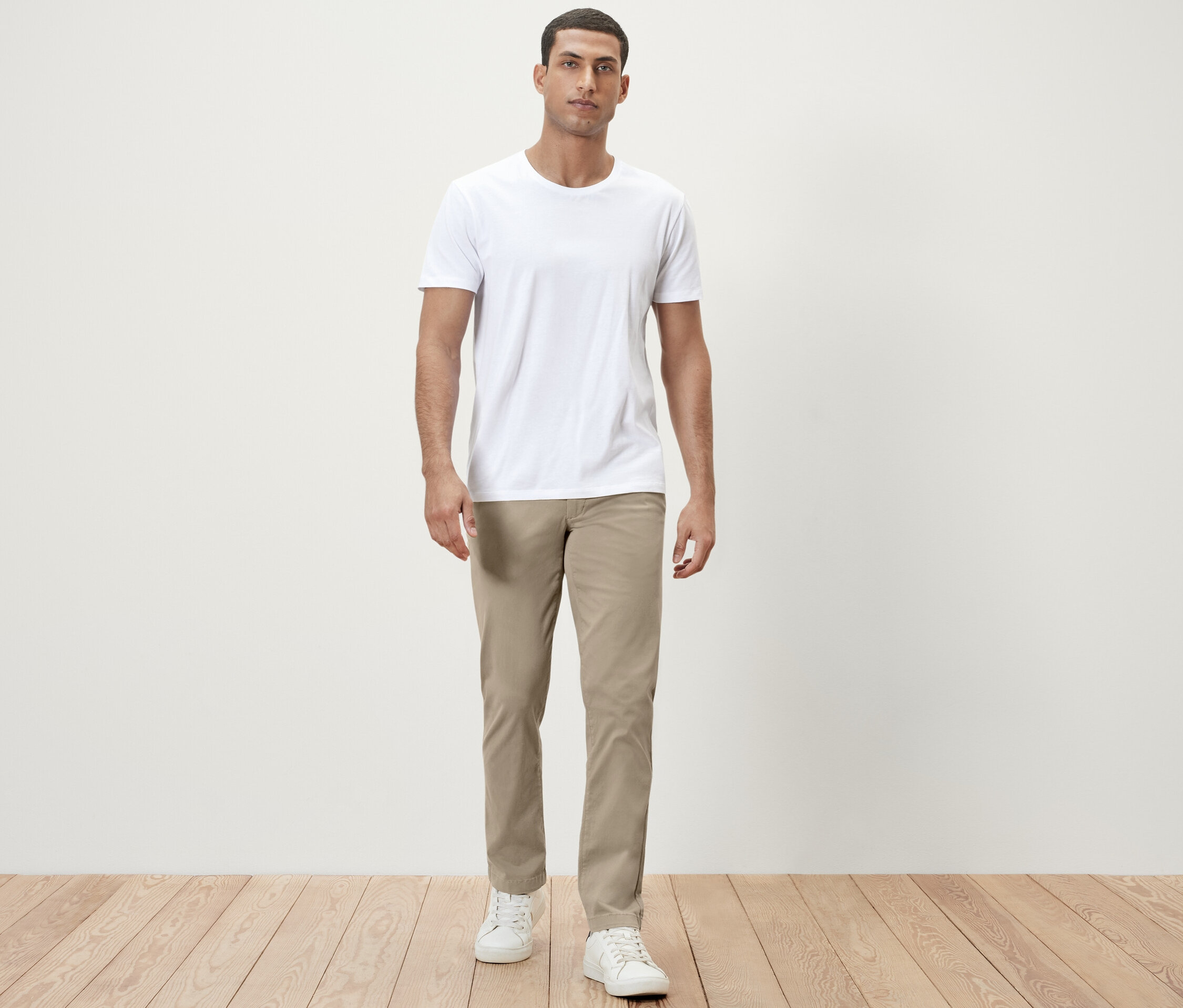 Muž stojí a má na sobě bílé tričko a béžové chino kalhoty.