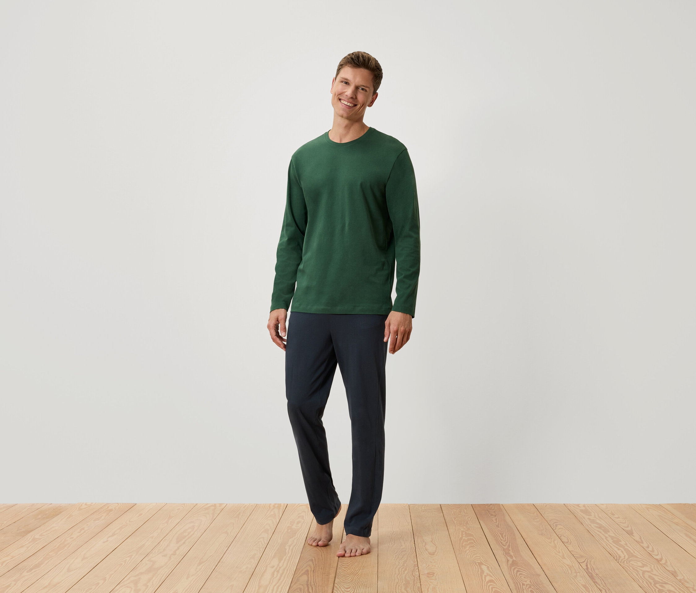 Muž stojí a má na sobě zelené tričko s dlouhým rukávem a tmavě modré kalhoty.