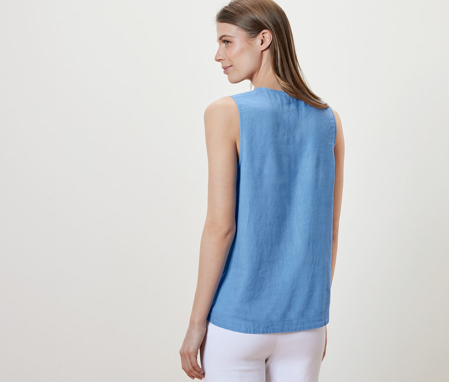 Žena stojí zády k fotoaparátu a má na sobě světle modrou lněnou halenku bez rukávů.