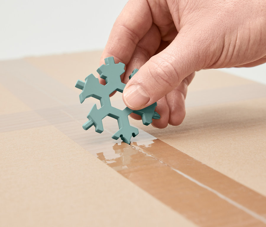 Ruka drží multifunkční klíč ve tvaru sněhové vločky a řeže s ním lepicí pásku na kartonové krabici.