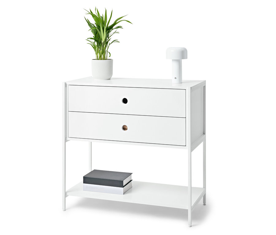 Bílý noční stolek se dvěma zásuvkami, lampou, rostlinou v květináči a knihami.