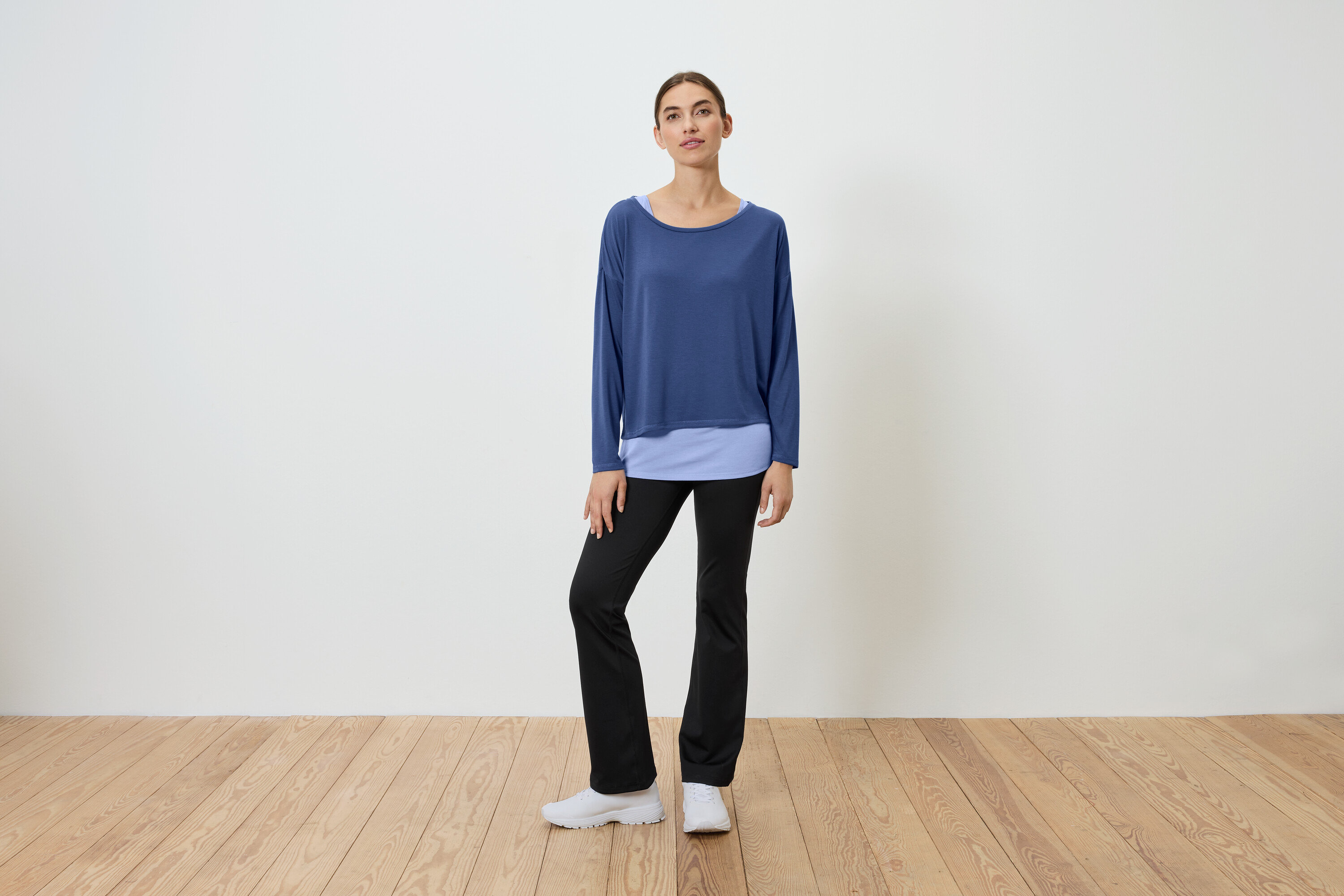Žena stojí na dřevěné podlaze a má na sobě modrý top a černé kalhoty.