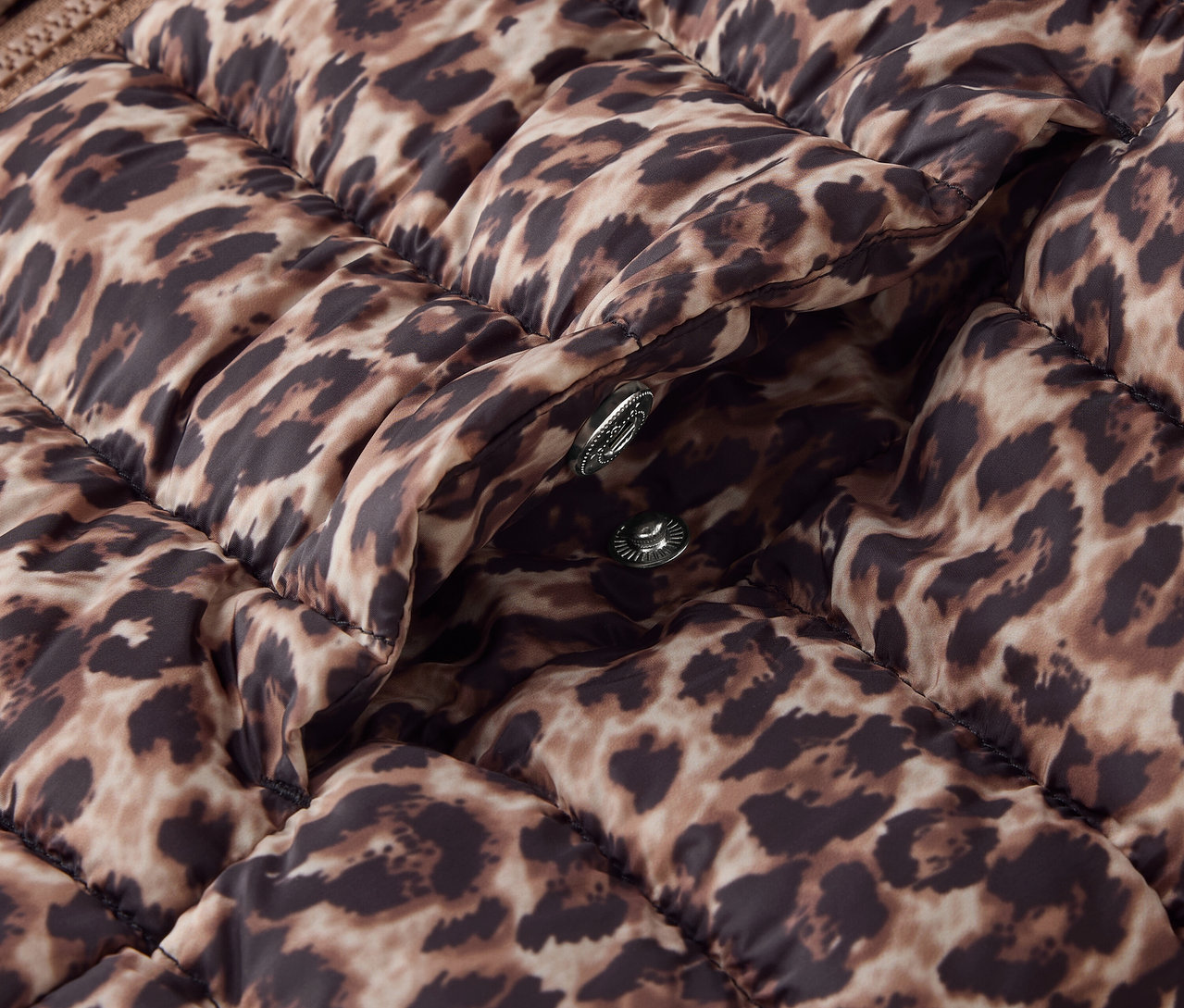 Detail hnědé prošívané bundy se zvířecím potiskem a stříbrnými patentkami.