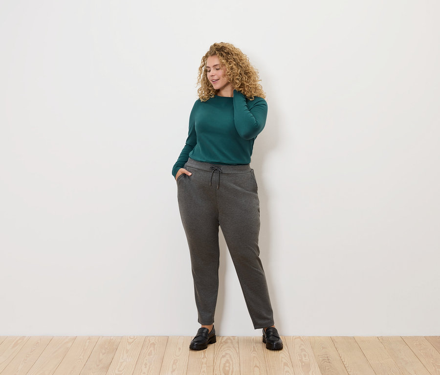 Žena s kudrnatými vlasy pózuje na světlém pozadí. Má na sobě zelenou blůzu s dlouhým rukávem a šedé tepláky.