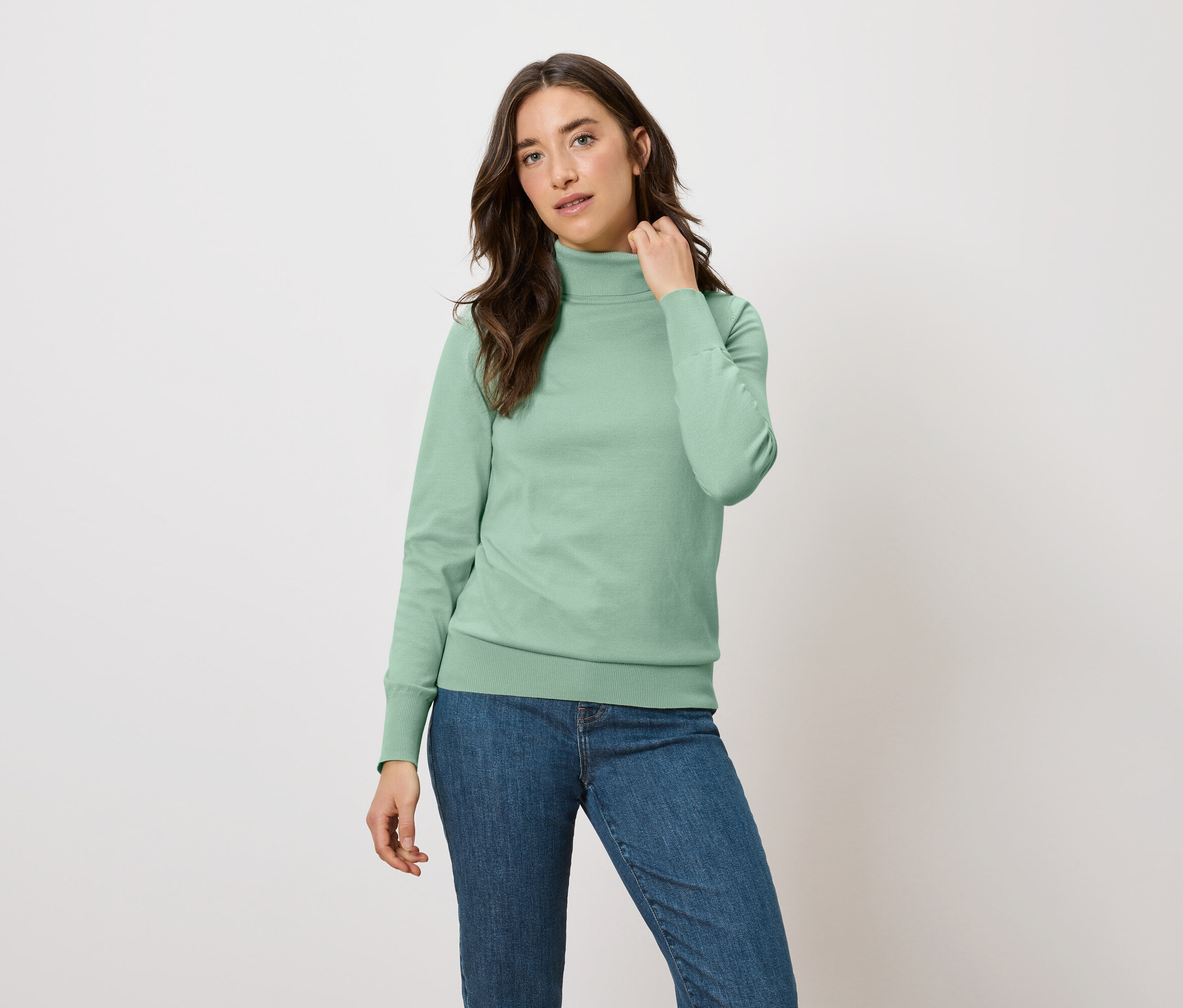 Žena pózuje v zelené mikině s rolákem a modrých džínách.