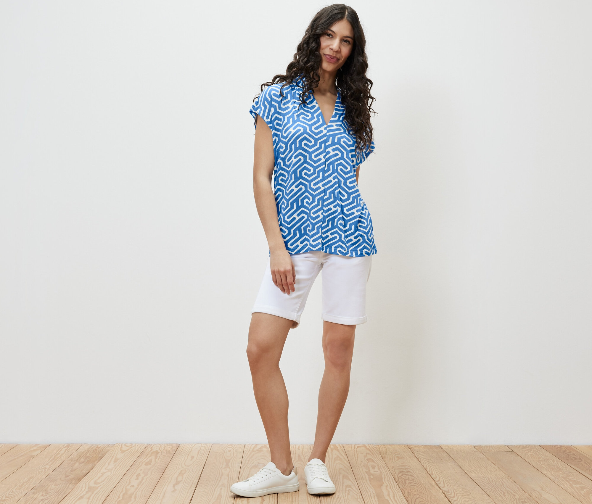 Žena stojí a má na sobě modré halenkové triko s výstřihem do V a bílé šortky.