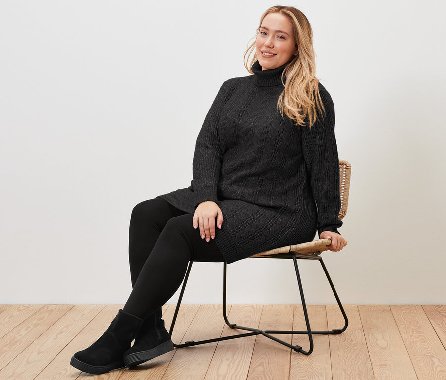 Žena sedí na židli a má na sobě dlouhý černý pletený svetr s copánkovým vzorem.