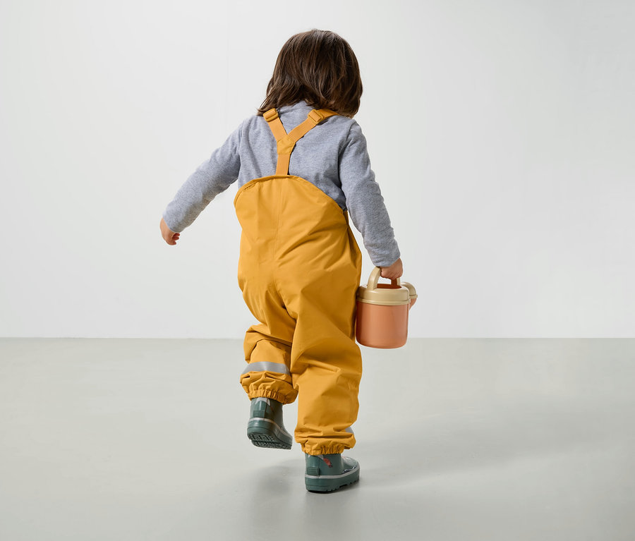 Chlapec v žlutých kalhotách s laclem a holínkách jde po podlaze a nese růžovou konev.